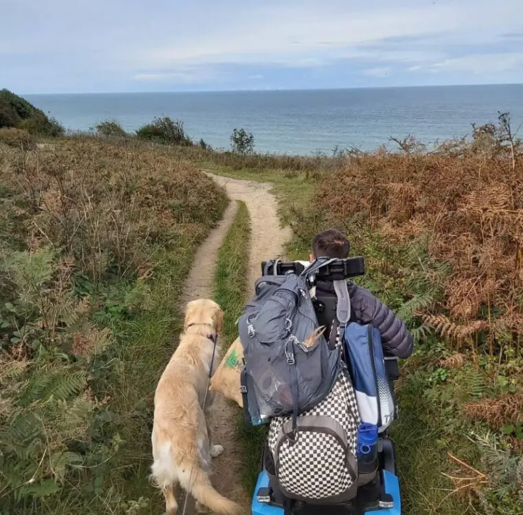 Martyn Sibley in his wheelchair holding dog Sunny on a lead walking along a path on the coast