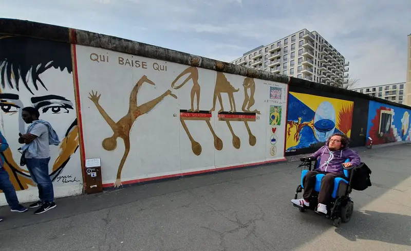 Derry Felton in his wheelchair beside the Berlin wall covered in artwork