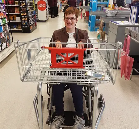 Lucy in a supermarket with a shopping basket on her wheelchair