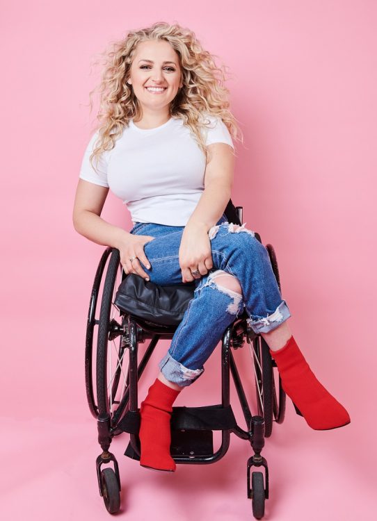 Ali Stroker in her wheelchair with a white t-shirt, jeans and red boots in front of a pink background