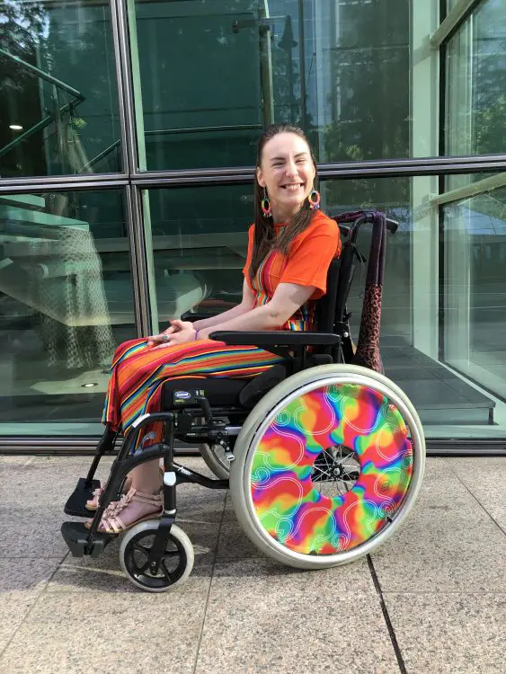 Rachel smiling and sitting her wheelchair in a bright orange outfit.