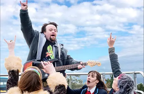 James Martin holding a guitar with his arm in the air at a music festival in the film Ups and Downs