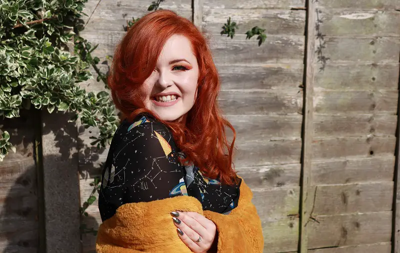 Lucy Edwards in front of a wooden fence with red hair wearing a black dress and yellow cardigan