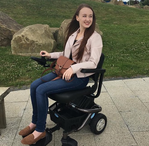Pippa in her wheelchair outside by a grassy bank wearing jeans and a pale pink jacket