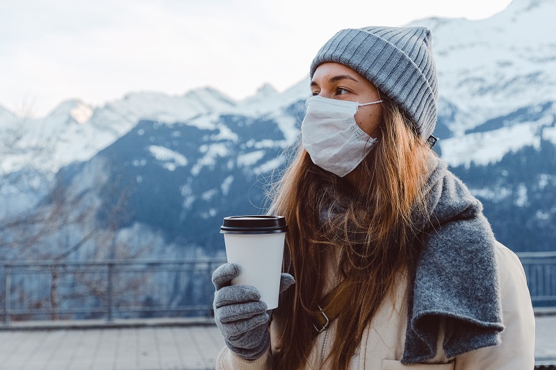 Woman in a grey scarf and cream coat stood in front of snow-covered mountains with a face mask on holiday a cup of coffee