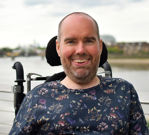 Toby Mildon in his wheelchair wearing a blue T-shirt with a floral pattern. He is sat in front of a river with buildings in the far distance