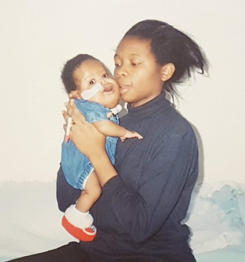 Olu as a baby in a denim babygrow with a feeding tube in his nose being held by his mother