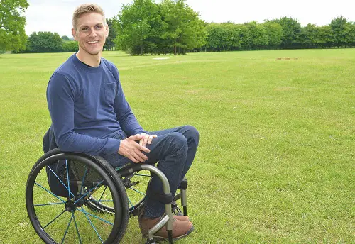 Steve Brown in a blue jumper and jeans sat in his wheelchair in a field with trees along the back