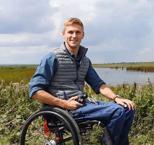 Steve Brown in a blue puffer jacket sat in his wheelchair in front of a lake and reeds