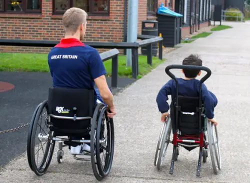 Steve Brown in his wheelchair next to a small boy in his wheelchair at a school wheeling away from the camera