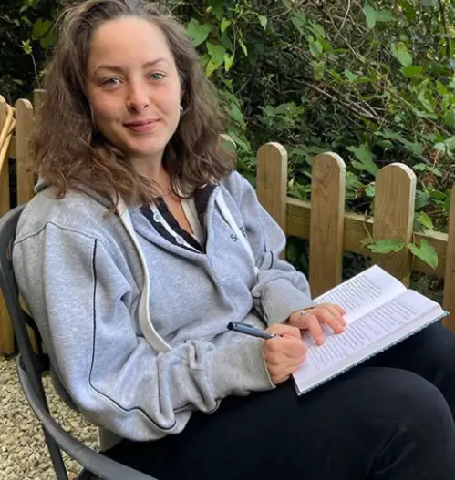 Catherine Shepherd in a grey hoddie and jeans sat on a grey chair in her garden with a notepad and pen