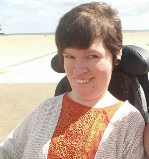 Gemma Orton in her wheelchair in a lacy orange top and white cardigain on a beach with the sea in the background