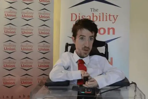 George Baker in his wheelchair with a white shirt and pink tie on in front of The Disability Union logos