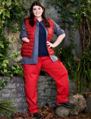  'Hollie Arnold wearing a red puffer jacket, blue top, red trousers and walking boots standing in front of a wall covered in green leaves ready for I'm a Celebrity'. 
