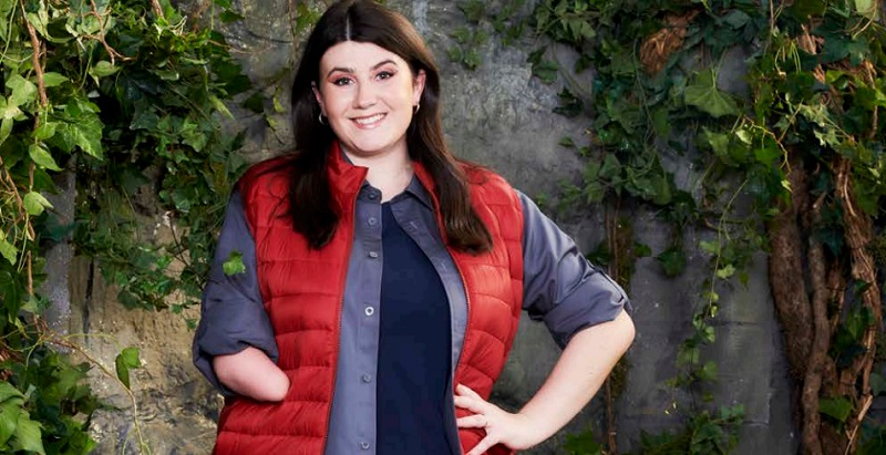 Hollie Arnold in a red puffer jacket and blue tops with her limb difference showing standing in front of a wall covered in leaves ready for I'm a Celebrity 2020