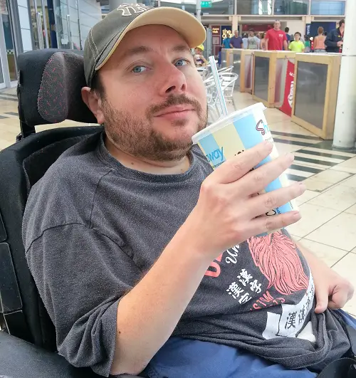 Sam Fairman in his wheelchair in a shopping centre wearing a grey t-shirt and blue trousers holding a takeaway drink
