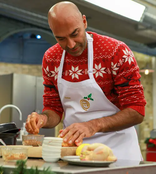 Blind presenter Amir Latif on Celebrity MasterChef Christmas special wearing a red Christmas jumper and white apron at a cooking station with ingredients