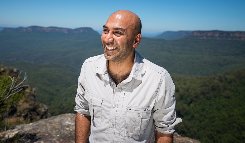 Blind presenter Amir Latif in a white short-sleeved shirt stood up high on a mountain with expanse of jungle behind him looking off to the left into the distance