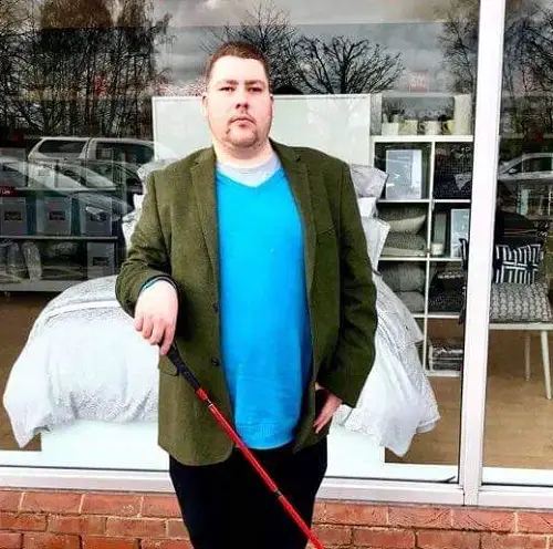Visually impaired blogger Luke Sam Snowdon wearing a bright blue T-shirt, green jacket, and black trousers with his red cane stood in front of a shop window