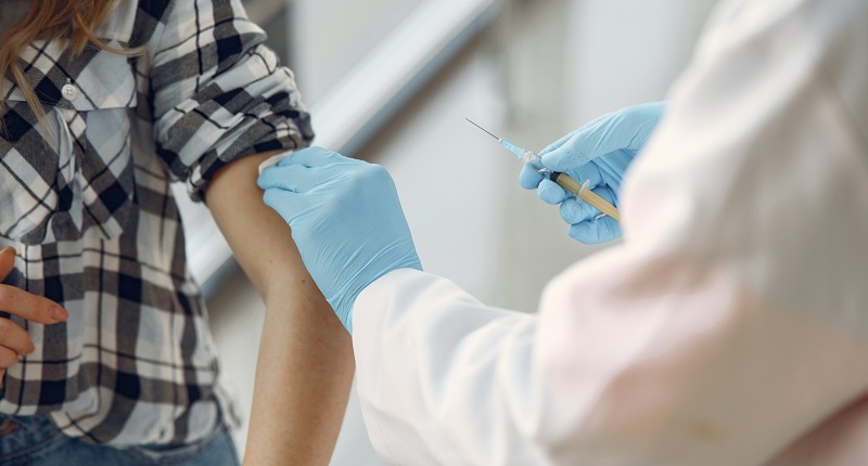 Woman i9n a black and white checked shirt about to be given a vaccine by a man in a white lab coat and blue plastic gloves