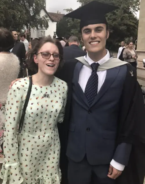 Sam and Ella at Sam's graduation with Sam wearing a dark grey suit and mortar board hat and Ella wearing a white dress with small yellow and purple flowers