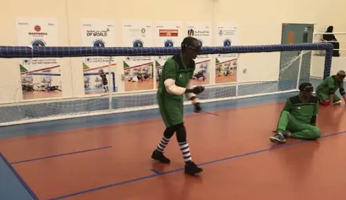 Yahya Siyad playing Goalball wearing a blindfold, green T-shirt and shorts, black and white socks and black shoes, in a sport's hall
