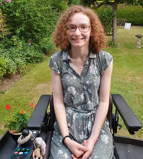 Hannah Deakin, smiling, sits in an electric wheelchair and wears a summery daisy patterned grey dress 
