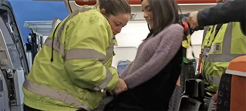 A lady is being manually lifted by two airport assistants into a airplane seat copy
