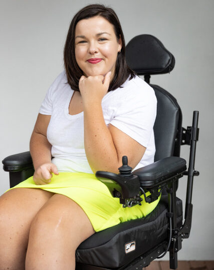 Peta Hooke in her wheelchair wearing a short yellow skirt and white T-shirt with short straight brown hair