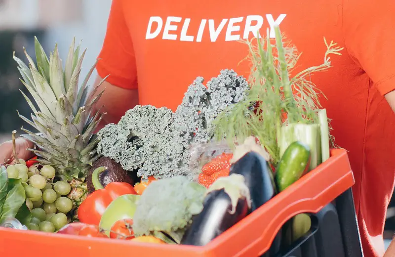 Man in an orange t-shirt with the word delivery on it delivering a box filled with food