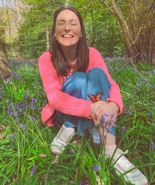 Rebecca is sitting in long grass, surrounded by flowers and is wearing a pink cardigan and jeans