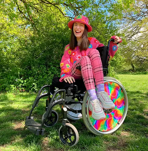 Rebecca is sitting sideways in her wheelchair, is wearing a pink outfit and is sat under a tree