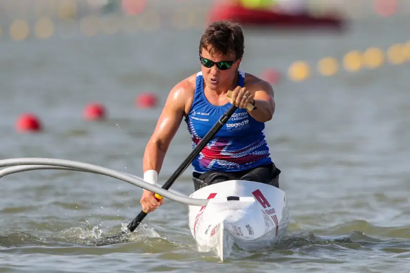 Emma Wiggs sitting in canoe on water with paddle down wearing sunglasses. Emma is wearing a blue vest with image of british flag