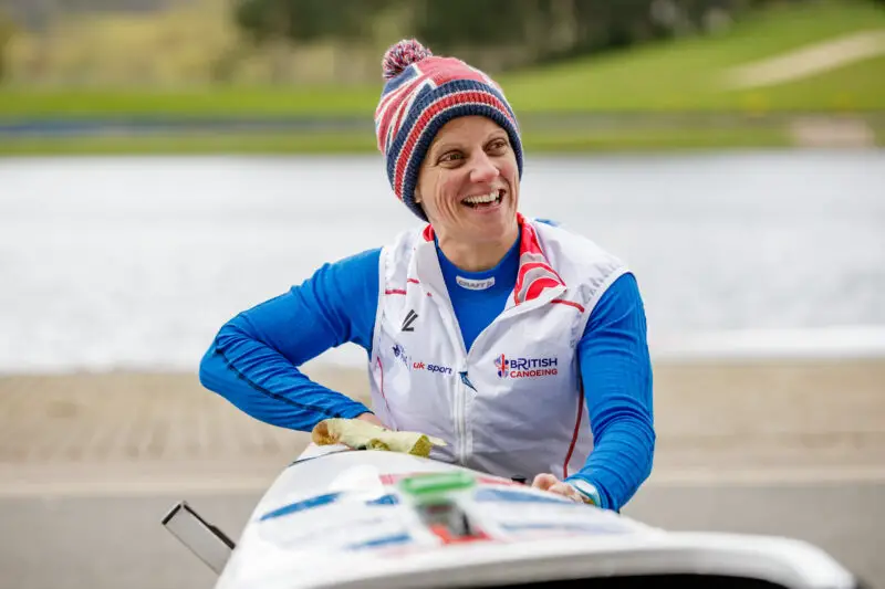 Emma Wiggs sitting in canoe smiling, wearing GB sports white jacket with hat design of British flag and pom pom at the back