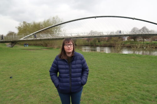 Holly-stood-on-some-grass-with-her-hands-in-her-pockets-she-is-wearing-a-blue-coat-and-blue-jeans-there-is-a-river-and-trees-in-the-background-and-a-tall-metal-bridge-in-the-distance-