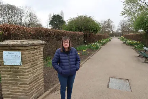 Holly-stood-on-a-stone-path-with-daffadils-and-brown-hedges-at-either-side-she-is-wearing-a-blue-padded-jacket-blue-jeans-and-grey-trainers-