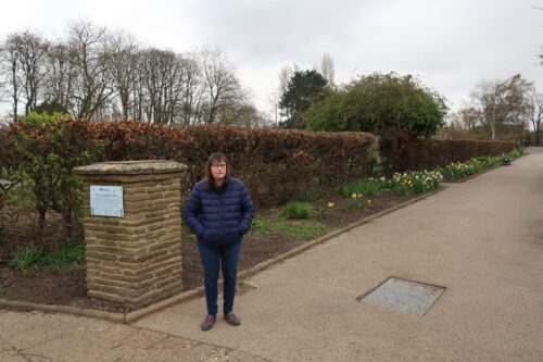 Holly-stood-on-a-stone-path-with-daffodils-and-brown-hedges-to-the-side-of-her-she-is-wearing-a-blue-padded-jacket-blue-jeans-and-grey-trainers-