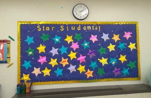 Classroom Star bulltein board. Image shows a blue board on a wall with the words 'star students' in gold letters at the top and lots of stars in green, yellow, pink and blue card to add student's names to