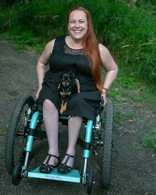 Nalani in her Mountain Trike all-terrain wheelchair in a wood with her dog on her lap