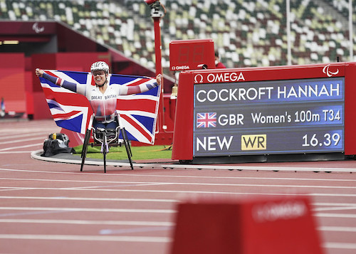 Hannah Cockroft holding union jack flag in her racing chair next to the digital board which shows T34 100 WR 16.39
