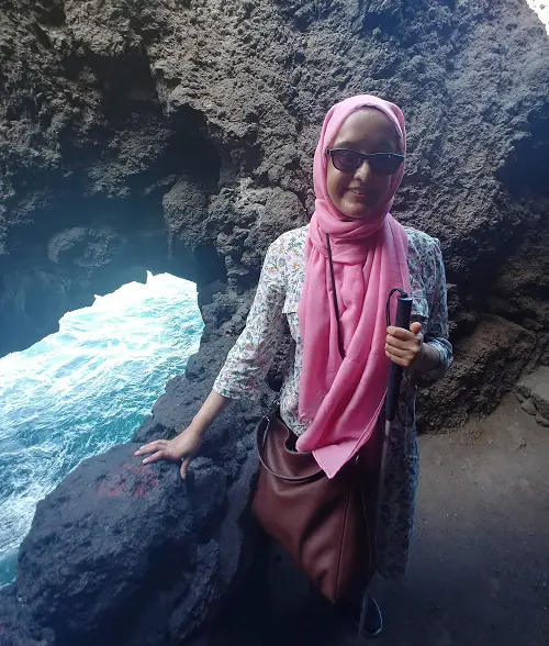 Nanjiba wearing glasses, a pink hijab, a denim jacket and dress standing next to water at a cove