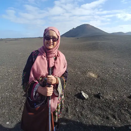 Nanjiba wearing glasses, a pink hijab and dress standing with her white cane in a dessert with a sand dune behind her