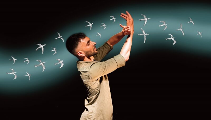 A man wearing a beige shirt and white jeans dancing in front of a dark wall with white birds flying across it - Little Murmurs at Imagine Children's Festival 2022 at Southbank Centre