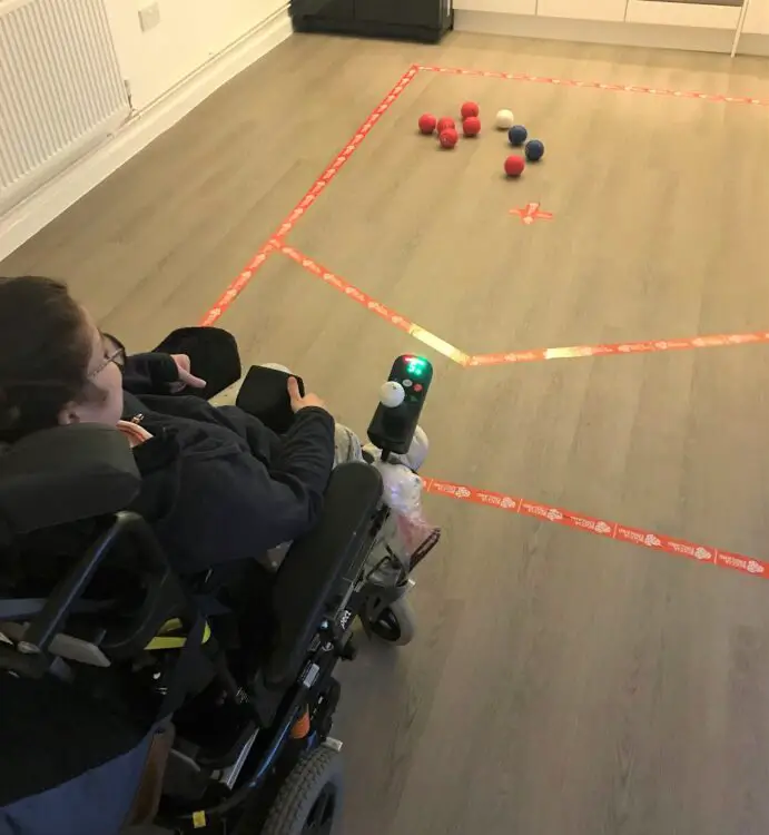 A girl in her power wheelchair playing boccia at home in her kitchen with a court marked on the floor using tape