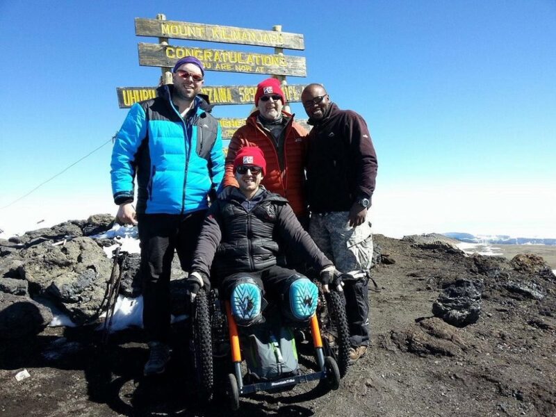 Four men, one in a Mountain Trike all-terrain wheelchair at the summit of Mount Kilimanjaro