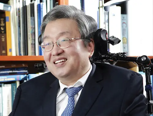 A close up of Lee Sang Mook in a wheelchair with short grey hair wearing a black suit, white shirt and blue tie and glasses in front of shelves of books