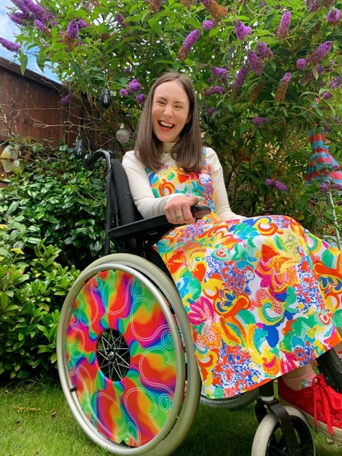 Rebecca with brown hair wear a colourful dress with a with polo, red shoes and a colourful beaded necklace. She sits in her wheelchair, with rainbow spoke guards in the garden surrounded by flowers.