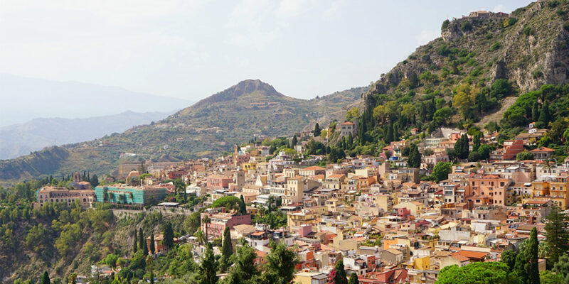 View from up high over Taormina, Sicily, showing colourful buildings in yellow, orange and red, amongst mountains covered with greenery