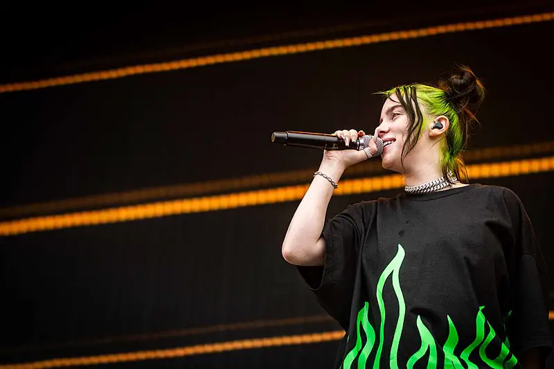 Billie Eilish wearing a black T-shirt with bright green flames at the bottom and her yellow and black hair in a ponytail on stage with a microphone held up to her mouth