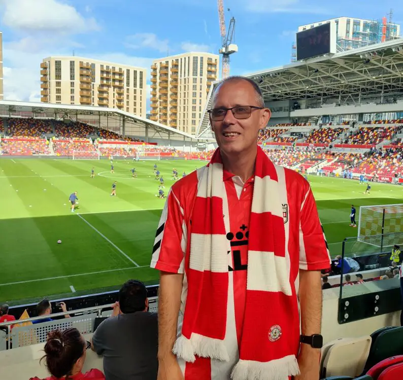 Gary Murphy at a football match standing in the stands with the pitch in the background wearing a red T-shirt and red and white striped scarf and wearing black glasses